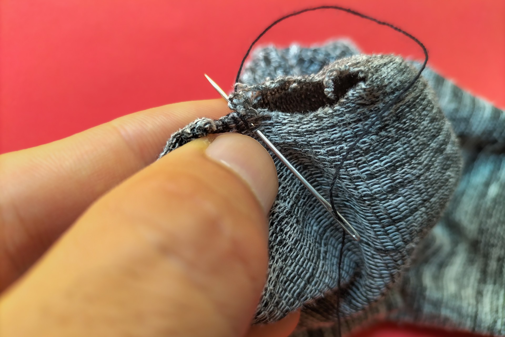 Hands of a white man sewing a hole in a striped gray sock with a sewing needle and black thread against a red background. Repair of clothes and ammunition, survival lifestyle, autonomy.